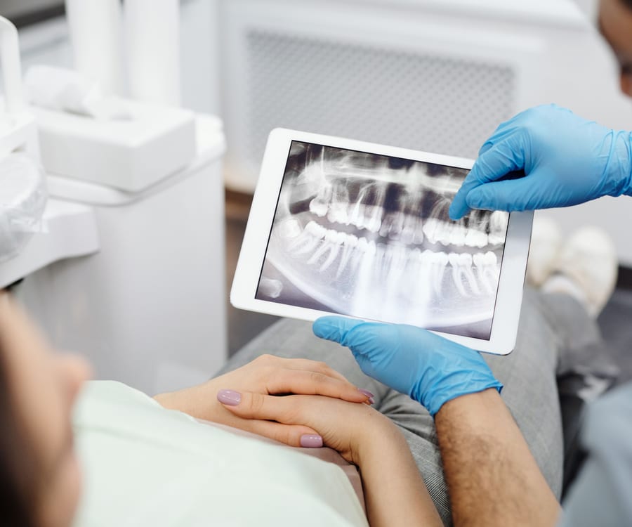 A gloved hand holds a tablet showing a panoramic dental x-ray to a person seated in a dental chair
